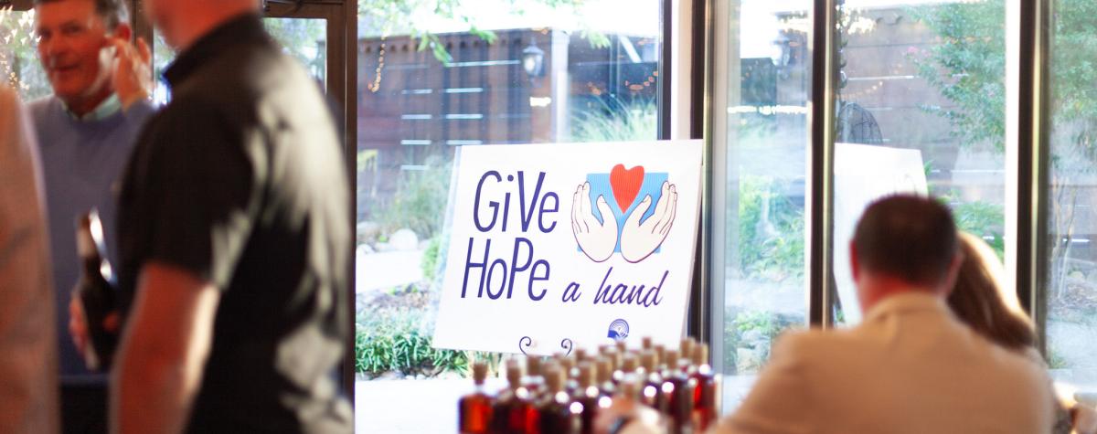 Sign that reads Give hope a hand with people in a well-lit room and bourbon bottles at a table.